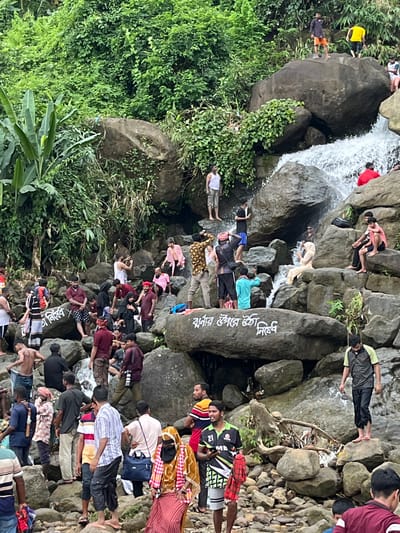 The image depicts a lively scene at a waterfall in Jaflong, Bangladesh. A diverse group of people are scattered across rocks and near the cascading water, engaged in various activities. Some individuals are standing on the rocks, observing the scenery, while others are seated. A few people appear to be wading in the water, enjoying the cool spray. Several individuals are dressed in casual clothing, including t-shirts, shorts, and traditional attire. Among the crowd, one person is seen taking a photograph, suggesting the scenic beauty of the location. The environment is characterized by lush green vegetation covering the rocky terrain and the presence of a significant waterfall. The atmosphere appears to be one of leisure and enjoyment, with many people gathered to experience the natural attraction.