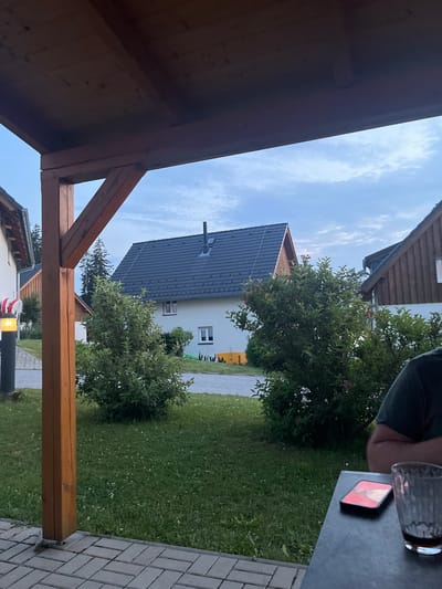 The image depicts an outdoor scene, likely a residential area in Milná, Czechia. In the foreground, a wooden support beam of a pergola is visible, with a tiled patio beneath it. Beyond the pergola, a lush green lawn stretches towards a street. Several houses with dark-tiled roofs and white facades are scattered across the landscape, interspersed with green trees and bushes. One house, more centrally located, has a prominent chimney. The sky is overcast with a soft, diffused light, suggesting it might be late afternoon or early evening. In the lower right corner of the image, a table is visible with a smartphone and a glass containing a dark liquid. A person's arm and shoulder are also present, indicating someone is seated at the table. A street lamp with a glowing orange light is positioned on the left side of the image, near one of the houses.