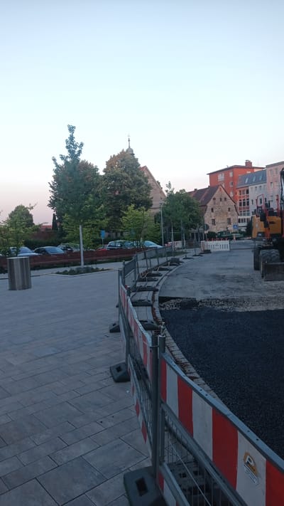 This image depicts a construction scene in Altdorf bei Nürnberg, Germany, likely a public area undergoing road or sidewalk repair. In the foreground, a temporary construction barrier with red and white striped panels separates the sidewalk from the work zone. The sidewalk is paved with large, grey rectangular stones. The construction zone itself is covered with dark gravel and asphalt, and includes a yellow excavator and other construction equipment. In the middle ground, several cars are parked along a street lined with trees and benches. In the background, several buildings are visible, including a church with a steeple and other residential or commercial structures. The sky is clear and appears to be either early morning or late evening due to the soft, diffused light. There are no people visible in the image.