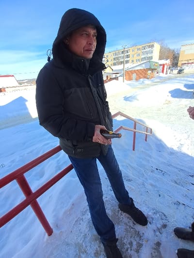 An outdoor scene in Bolshaya Elhovka, Russia, depicts an adult male standing on a snow and ice-covered ground under a clear blue sky. The male is positioned slightly right of the camera's center, looking towards the right, and is wearing a dark hooded puffer jacket with the hood up, blue jeans, and dark boots. He holds a dark object with a yellow/orange tip in his right hand; a ring is visible on his left ring finger. A red metal handrail is present in the lower left and center foreground. Partially visible on the far right is the lower leg and dark boot of another individual. The background features a multi-story, light-colored apartment building in the center, flanked by smaller commercial or residential structures. The terrain is covered in a substantial layer of snow and ice, with patches of cleared ground. Leafless trees are visible in the distance, and a snow-covered road or parking area with a dark vehicle is also discernible.