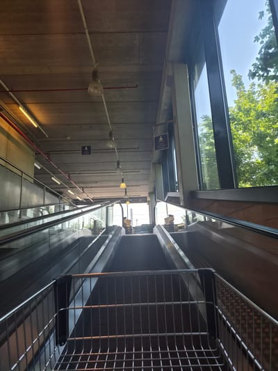 This image shows an indoor escalator with a shopping cart in the foreground. The escalator is made of metal and glass, and it is moving upwards. There are lights on the ceiling, and a large window on the right side of the image looks out onto trees and a blue sky. The location is identified as Sanem, Luxembourg. There are no people visible in the image. The scene appears to be a public space, possibly a shopping mall or a transportation hub. The image is taken from a low angle, looking up the escalator.