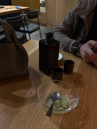 This image captures an indoor dining scene, likely within a restaurant or bar in La Purísima, Mexico. The perspective is a close-up on a light-colored wooden table with a distinct grain. On the left, a light brown/taupe textured handbag with a gathered top handle rests on the table. Centrally, a tall, rectangular, dark brown or black ceramic bottle with a narrow, slightly undulating neck is positioned. To its right are two small, dark brown/black ceramic cups and a small, white circular dish partially visible behind them. In the foreground right, a clear glass bowl contains remnants of a light green, lumpy substance, possibly a dessert or appetizer, and a small, metallic spoon with a patterned handle lies within the bowl. To the far right, the lower torso and left arm of a person are visible; they are wearing a light brown or grey long-sleeved jacket or shirt and multiple wristbands or a watch. This individual's hands are occupied with a dark-colored mobile phone. The background is softly blurred, showing other wooden tables and dark-colored chairs, typical of a restaurant setting, under dim, warm lighting.