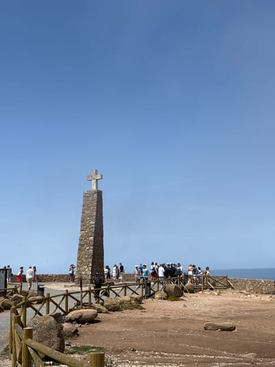 The image shows the monument at Cabo da Roca, the westernmost point of mainland Europe, located in the municipality of Colares, Portugal. A tall, stone monument topped with a cross stands prominently against a clear blue sky. A group of tourists is gathered around the monument, some standing on a paved area behind a wooden fence, and others further back on a stone wall overlooking the ocean. The ground in the foreground is a mix of dirt, rocks, and gravel, with more wooden fencing. The ocean is visible in the background. The scene depicts a popular tourist destination on a sunny day.
