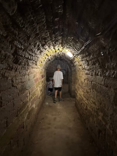 Two individuals, an adult and a child, are walking through a dimly lit, arched stone tunnel. The tunnel walls are constructed from rough-hewn, large stone blocks, some of which appear to be reddish-brown and others a lighter, sandy color, with patches of moss or algae. The ceiling of the tunnel is also made of stone and appears roughly shaped. Two modern, rectangular light fixtures are visible on the ceiling, providing illumination. The floor of the tunnel is dirt or gravel. The adult, wearing a white t-shirt, black shorts, and a light-colored baseball cap, is walking ahead of the child, who is wearing a dark t-shirt, shorts, and a blue baseball cap. The image was taken in Posyolok Imeni Morozova, Russia.