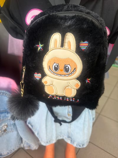 A person wearing a black shirt with a pink circular design and a black fuzzy backpack with a cartoon bunny embroidery is shown from the chest down. The backpack has colorful embroidered stars and hearts. A dark furry pom-pom is attached to the side of the backpack by a gold chain. The person is also wearing a light blue denim skirt. The background is out of focus and appears to be indoors, possibly a retail store, with some merchandise visible. The scene is located in the city of Ravno pole, Bulgaria.