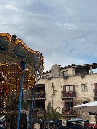 A vintage carousel is brightly lit against a building and a sky filled with clouds. The carousel's ornate detail is visible, with many lit bulbs accentuating its curves and artistry. The multi-story building behind it shows aged plaster and climbing vines. Windows with shutters and small balconies add to the charm. There are people behind and to the right side of the carousel.