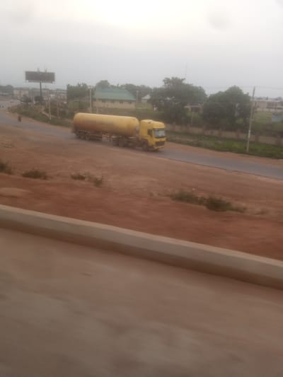 An over-exposed, hazy, daytime, eye-level, full shot depicts a yellow tanker truck driving on a paved road. The truck is positioned on the right side of the frame, angled away from the viewer and slightly to the left. The tanker is cylindrical and light yellow. The front of the truck, the cabin, is dark yellow with a black grill and white headlights. The truck has multiple wheels. The road is dark gray and appears to be a highway with no lane markings visible. The road is bordered on the left by a reddish-brown dirt embankment with some sparse vegetation. On the right side of the road, there are more trees and some buildings, including a house with a green roof and a billboard. The sky is overcast and a light gray color, suggesting either dawn, dusk, or a cloudy day. The image appears to be taken from a moving vehicle, as indicated by the motion blur in the foreground, which shows a light brown surface.