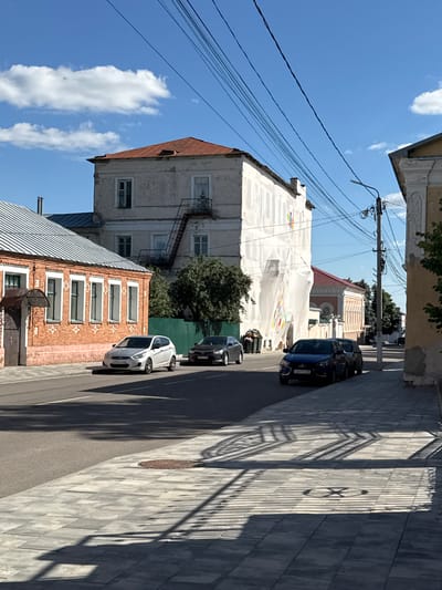The image captures a street scene in the city of Yelets, Russia, under a clear blue sky with scattered white clouds. The foreground features a sidewalk paved with gray rectangular tiles, casting long shadows from an unseen overhead structure. A manhole cover with a circular pattern is visible on the sidewalk. The street itself is paved with asphalt and shows a white lane marking.
Several cars are parked and driving on the street. A white hatchback is in the left lane, followed by a dark sedan. Further down the street, a dark blue sedan is parked, with another dark car partially visible behind it.
Buildings line both sides of the street. On the left, a single-story brick building with a metal roof has multiple windows with decorative white trim. Adjacent to it, a taller, older building with a weathered facade and a rusty metal roof is covered in part by white sheeting, with an external metal staircase attached. Colorful decorations, possibly flags or banners, are visible on the white sheeting. Further down this side of the street, a building with an orange-tiled roof and cream-colored walls is visible.
On the right side of the street, a building with a pale yellow facade and ornate architectural details is partially visible, with a streetlamp and utility pole in front of it. The overall impression is a somewhat aged urban environment with ongoing renovations or improvements suggested by the sheeting on one building.
