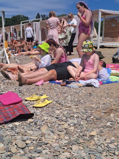 The image depicts a crowded beach scene in Dzhubga, Russia, under a clear blue sky. Numerous people are sunbathing and relaxing on a pebble beach. In the foreground, a child wearing a bright green hat and a pink swimsuit is eating. Next to the child, another young girl with long purple braided hair is also in a pink swimsuit. Several adults are visible in the background, some sitting and others standing. Beach chairs, towels, and bags are scattered around. The environment is characterized by the rough texture of the pebbles covering the ground and the presence of beach cabins in the distance.
