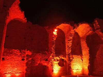The image depicts the interior of the Romanesque church of San Francesco in Alghero, Italy, illuminated by dramatic red and orange lights at night. The scene focuses on the ancient stone walls and arched openings, casting deep shadows and highlighting the rough texture of the masonry. The lighting creates an atmospheric and somewhat stark ambiance. There are no people or visible objects other than the architectural elements of the ruins themselves. The environment is dark, with the blackness of the night sky visible above the structure.