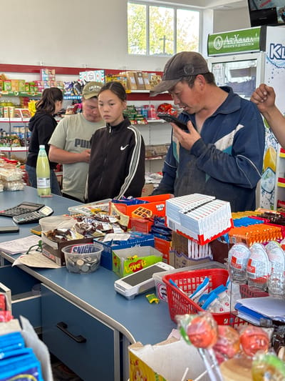 The image captures a busy scene inside a small grocery store or market in Kyzyl-Suu, Kyrgyzstan, with the camera oriented vertically. The setting features a blue counter serving as a point of transaction or display.
In the foreground, a man in a dark blue jacket and a baseball cap is intently looking at a black smartphone he holds. His hand is also visible higher up, possibly another person's arm, suggesting interaction or work at the counter. Behind him, a young woman in a black track jacket with white stripes looks towards the left, appearing to be observing or waiting. Further back, another man in a light-colored t-shirt with a "Puma" logo and a cap that says "COLUMBIA ATHLETIC" on the side is visible, along with a woman in a black top who is partially obscured.
The counter is laden with various products and store items. On the right, there are numerous stacks of "Kinder" branded items, boxes of "Waffle Cake," and other packaged snacks. A clear plastic container filled with coins labeled "SNICKERS" is also present, alongside a calculator and scattered papers, including receipts. Lollipops on sticks are displayed prominently, partially obscuring other items. Further down the counter, a light green bottled beverage is visible.
In the background, shelves are stocked with various goods, including a "HUGGIES" branded product. A refrigerator with "АлХалал КОЛБАСЫ И ДЕЛИКАТЕСЫ" (AlHalal Sausages and Delicatessen) text is noticeable, indicating specific product offerings. A window allows natural light into the store, showing trees outside, suggesting it is daytime. Other visible text includes "Love is..." and "KL" on various product displays. The overall scene depicts a typical commercial exchange or moment of activity within a local shop.