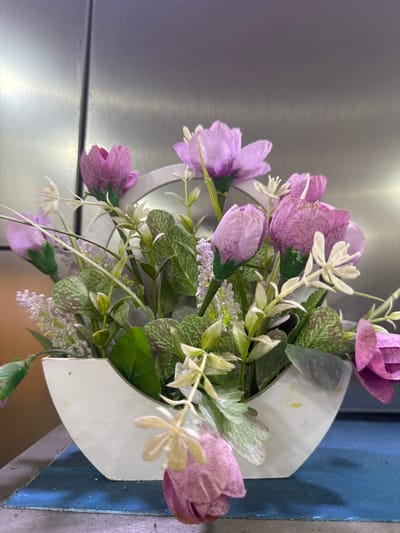 A floral arrangement in a white, U-shaped vase sits on a blue surface. The vase contains artificial flowers and greenery, predominantly in shades of pink and purple, with some lighter accents. The background is a metallic, brushed surface. The scene appears to be indoors, possibly a domestic or commercial setting in Shofirkon, Uzbekistan.