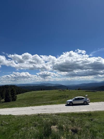 The image depicts a scenic mountain landscape with a silver sedan parked on the side of a paved road. The car's trunk is open, suggesting the occupants might be engaged in an activity such as sightseeing or picnicking. The road curves into the distance, bordered by lush green grass and rolling hills. In the background, a dense forest of evergreen trees stretches across the slopes, leading to a series of layered mountain ranges under a vast, blue sky dotted with white, fluffy clouds. The lighting indicates it is daytime, likely late morning or early afternoon, with the sun casting shadows. The overall scene is serene and evokes a sense of natural beauty and tranquility. The location is identified as Cherna Mesta, Bulgaria.