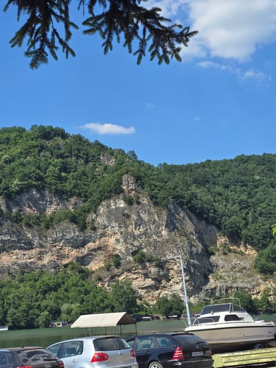 In the city of Divic, Bosnia and Herzegovina, this image captures a scenic view of a rocky cliff face covered in lush green trees, overlooking a calm body of water. The sky is a clear blue with a few scattered white clouds. In the foreground, several cars are parked near the water's edge, and a white boat rests on a trailer. A white canopy structure is also visible. The overall impression is one of a peaceful, natural setting.