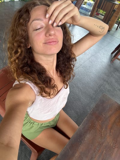 A woman with long, curly brown hair is depicted reclining in an outdoor or semi-outdoor setting in Du Già Commune, Vietnam. She is wearing a white t-shirt and green shorts. Her eyes are closed, and her left hand is resting on her forehead, suggesting a moment of relaxation or rest. A distinct outline tattoo, possibly of a creature like a bull, with four small dots below it, is visible on her left arm. Her nails appear manicured.

The background features a grey tiled floor and dark brown wooden furniture, including a table and what appears to be a plastic chair. Wooden structural elements or railings are visible, and in the distant background, a blue and white patterned area, possibly water or a pool, can be seen. The scene is brightly lit by natural light, indicating it is daytime. A partial label, green and white, with the word "LAGER" is faintly visible near her head. The overall impression is one of calm and leisure in a tropical or semi-tropical environment.
