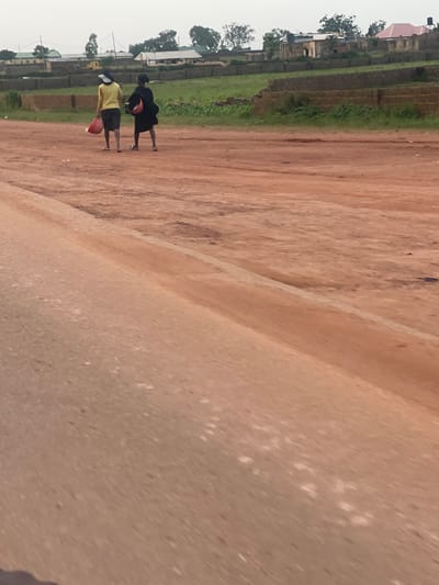Two people are standing on a dirt road. One person is wearing a yellow shirt and dark shorts, and the other is wearing a black dress. Both individuals are holding red buckets. Behind them, there is a grassy area and a row of buildings with red roofs. The sky appears overcast, suggesting it might be a cloudy day. The overall scene depicts a rural or semi-urban environment in Rayfield, Nigeria. The people seem to be engaged in carrying water or some other task, as indicated by the buckets they hold. There is no visible text in the image.