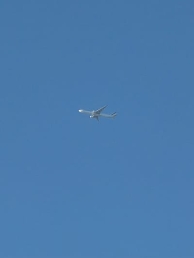 An airplane is captured flying across a clear blue sky. The aircraft, a commercial jetliner with two engines mounted under its wings, is seen from a slightly below and side angle. The plane appears to be white with a blue tail. There are no other objects or structures visible in the frame, indicating an aerial perspective high above any land-based features. The location is stated to be Riverhead, United States of America, though no specific landmarks or environmental details within Riverhead are visible to confirm this.