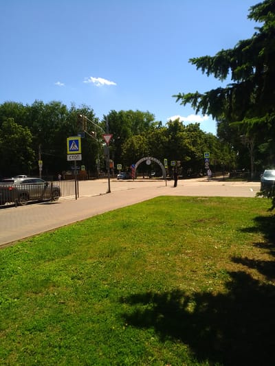 The image depicts a street scene in Otradny, Russia, on a clear, sunny day. In the foreground, a well-maintained lawn occupies the lower right and a portion of the lower left of the frame. A paved sidewalk runs along the left side of the image, separating the lawn from the street.
The street itself features pedestrian crossings and traffic lights. Several traffic signs are visible, including a "Stop" sign and a pedestrian crossing sign with a blue background and a white figure. A dark-colored car is parked on the left side of the street, behind a metal fence.
In the background, a dense line of lush green trees forms a backdrop to the scene. Across the street, an arched gateway with lettering is visible, suggesting an entrance to a park or public space. Two individuals are present in the midground: one man is walking across the street, while another, dressed in dark clothing, stands near the sidewalk. The sky above is a clear, bright blue with a few scattered white clouds. The overall impression is one of a peaceful urban environment during daylight hours.