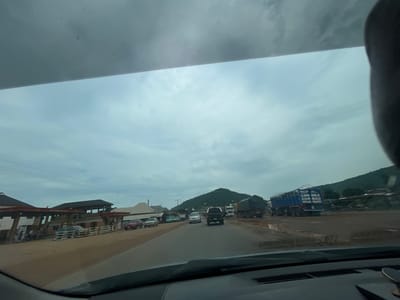 The image depicts a view from inside a vehicle, looking out through the windshield and partly obscured by the car's dashboard. The scene is a road with several vehicles, including cars and trucks, suggesting moderate traffic. On the left side of the road, there are buildings that appear to be commercial establishments, possibly a gas station with multiple pumps visible. Further down the road, a large blue truck with a cage-like trailer is prominent, as are other large trucks, some carrying what appear to be loads covered with tarpaulins. In the background, lush green hills rise against a cloudy, overcast sky. The road surface appears to be asphalt, with some areas showing signs of mud or puddles, indicating recent rain or a damp environment. The overall atmosphere is that of a semi-urban or roadside setting in a tropical or subtropical region, consistent with Koton Karifi, Nigeria. No individuals are clearly discernible in the image.