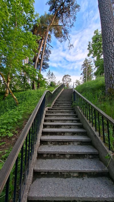 A long, outdoor stone staircase with dark metal railings leads upwards on a green, grassy slope dotted with trees under a partly cloudy blue sky. The staircase is made of concrete with visible aggregate. The railing on the left is wooden on top of the metal. Lush green grass and various trees, including tall pines with rough bark and deciduous trees with bright green leaves, surround the staircase. The lighting suggests it is either morning or late afternoon. The scene is set in Keila-Joa alevik, Estonia.
