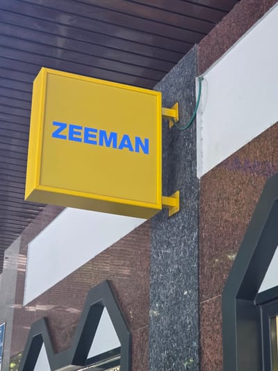 A yellow square sign with blue lettering that reads "ZEEMAN" is mounted on a building facade. The sign is attached to the building by two yellow brackets. The facade features a combination of granite and white marble panels. A dark brown wooden soffit is visible above the sign. A green wire extends from the sign to the building. A portion of a black angular architectural element is present in the lower right of the image. The overall scene suggests an exterior view of a commercial establishment. The location is identified as Collado Villalba, Spain.