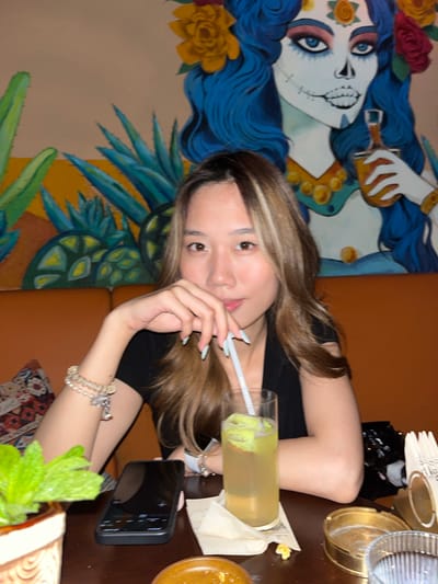 The media file presents a young woman in an indoor setting, likely a themed restaurant or bar, with a location context of Longta, China. The image is rotated 90 degrees clockwise.
The subject is a young woman with long, light brown hair featuring blonde highlights, wearing a black top. She looks directly at the camera with a calm, neutral expression. Her right hand holds a white straw from a tall glass containing a yellowish drink with green garnishes. Her fingernails are painted a light blue or mint green. On her right wrist, she wears multiple bracelets, including one with pearls and another with a cross charm.
She is seated at a dark wooden table, where a black smartphone rests. Other objects partially visible on the table include paper napkins and a gold-colored plate or coaster. A leafy green plant is in the foreground.
The background features a colorful mural on the wall, depicting a stylized female face with vibrant blue hair, reminiscent of a Calavera or Day of the Dead skull. This artwork includes prominent blue eyes, yellow roses, and blue agave-like plants, suggesting a Mexican or Latin American theme. The seating appears to be an orange-brown upholstered booth or bench, with a patterned rug visible beneath the table. The ambiance suggests a relaxed social gathering. No discernible text is visible in the image.