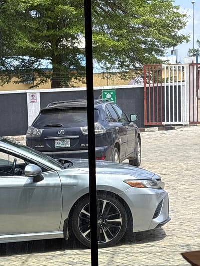 The image shows a parked silver sedan in the foreground, with its front end partially visible. Behind it, a dark blue Lexus SUV is parked. The SUV has a license plate that reads "SMK 396HS". To the left of the SUV, a "No Parking" sign is visible. To the right of the SUV, a fence with red and white bars separates the parking area from an unknown area. The ground is paved with light-colored interlocking bricks. In the background, a large green tree with dense foliage dominates the upper part of the image, casting shadows. Behind the tree and fence, there are buildings with yellow and white walls. The sky is visible in the upper right corner, appearing bright and possibly overcast. The scene appears to be a street or parking lot in Gwaska, Nigeria.