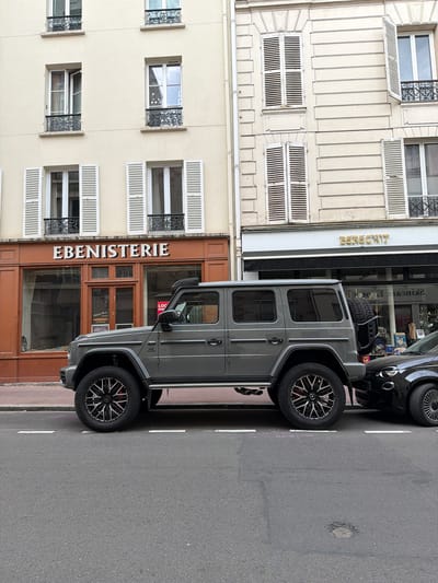 The image captures a street scene in Levallois-Perret, France. A gray Mercedes-Benz G-Class SUV is parked on the street, close to the curb. The car is positioned in front of a storefront with the word "EBENISTERIE" displayed above its entrance. The building adjacent to the EBENISTERIE has a beige facade with multiple windows, some featuring white shutters and decorative ironwork. A partial view of a black vehicle is visible behind the G-Class SUV. The street is paved with asphalt and marked with white lane dividers.