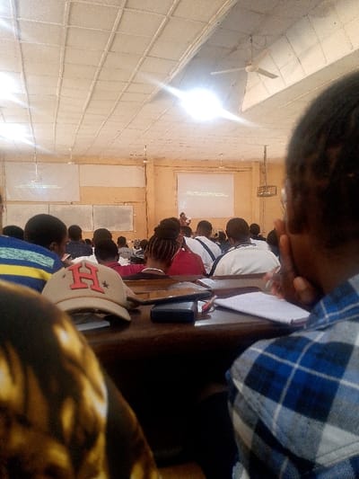 The image depicts a lecture hall or large classroom within the city of Alabata, Nigeria. The room features light-colored walls, possibly beige or cream, and a grid-paneled ceiling with recessed lighting and at least two visible white ceiling fans. Bright overhead lights illuminate the space, with one causing a significant lens flare.

Approximately two dozen individuals, primarily young adults, are seated in rows at dark wooden desks, facing the front of the room. In the foreground right, a person wearing a blue and white plaid shirt is observed from behind, holding a pen over a notebook placed on a desk. A black smartphone and a red-capped pen lie nearby on the same desk. To the foreground left, a tan baseball cap with a red 'H' logo rests on an adjacent wooden desk, partially obscuring another attendee wearing a brown and gold patterned top.

Mid-ground attendees include individuals with braided hair in a red top and others in white tops with dark detailing. At the front of the room, a person stands near a central projection. Two rectangular screens or whiteboards display projected text; the right screen clearly shows "THIS ONE IS OUR NATIONAL" above "STUDENT INDUSTRIAL WORK EXPERIENCE SCHEME." The assembled group is oriented towards the front, indicative of attending a lecture or presentation.