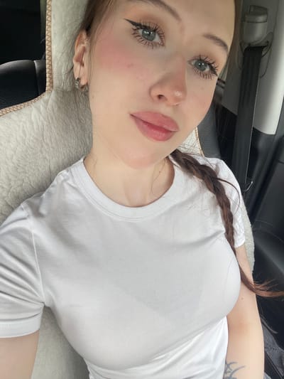 The image shows a young woman with light skin and light blue eyes, wearing a white t-shirt. She has her hair in a braid and is wearing subtle makeup. The woman is sitting in what appears to be a car, as indicated by the seatbelt visible on the right side of the frame and the car seat upholstery on the left. There is no information to suggest this image is located in Gavrilovka, Russia. The background is out of focus, but the interior of a vehicle is discernible.