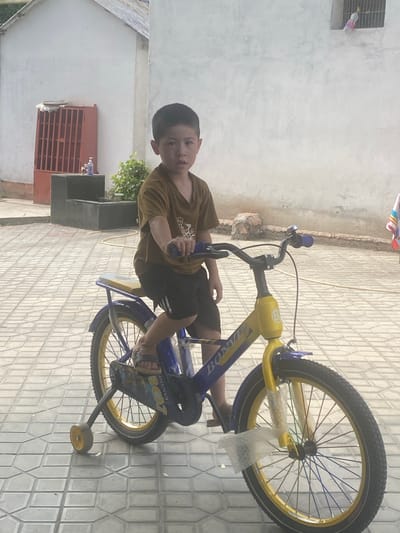 A young boy is sitting on a blue and yellow bicycle with training wheels, on a tiled patio. He is wearing a brown t-shirt and black shorts. The bicycle has a bell on the handlebars and has some protective plastic wrapping on the front. In the background, there is a white building with a red metal grate on one side. There is also some greenery and a rocky outcrop. A small flag is visible on the right side of the image. This scene appears to be located in the city of Kokand, Uzbekistan.