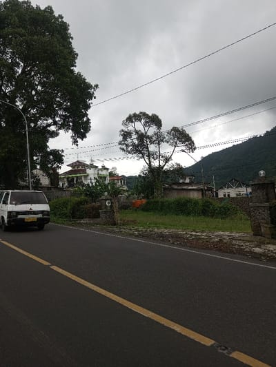 The image depicts a roadside scene in Kodarapalli, India, under a uniform overcast sky. In the foreground, an asphalt road with a solid yellow center line extends from the lower left to the upper right. A white Mitsubishi van, positioned on the left side of the road, faces right with its headlights visible and a yellow license plate affixed to the front bumper.
To the right of the road, an area of overgrown green vegetation precedes a pair of stone pillars, possibly marking an entrance, each topped with a spherical, light-colored fixture. Behind the vegetation on the left, a large tree with dense green foliage stands. Further back, a multi-story building with white walls, a reddish-brown tiled roof, and green trim features several architectural elements resembling small spires or decorative finials on its roof. Another tree with a more open canopy is situated to the right of this building.
Multiple utility poles with overhead power lines traverse the scene at various elevations. In the mid-distance, behind the entrance pillars, an orange water tank is visible near a low-rise structure, and a partially constructed or damaged white building with exposed framework is present on the far right. The background is dominated by a dense, green mountainous or hilly landscape. No human figures are present.