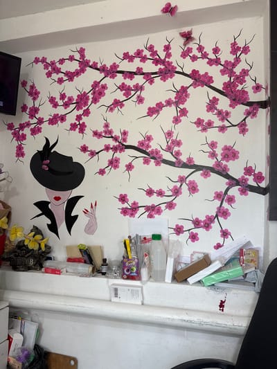 The image depicts a wall mural of a cherry blossom tree with pink flowers and two pink butterflies. A stylized portrait of a woman in a black hat and jacket, with red lips and red fingernails, is painted on the wall below the tree. The wall is white and appears to be in a room with shelves and various items placed on them. The items include bottles, containers, boxes, papers, and what appear to be art supplies. The scene is lit with artificial light. The overall impression is of a decorated interior space. There is no indication of the city of Aleksandrovskiy, Russia within the image itself.