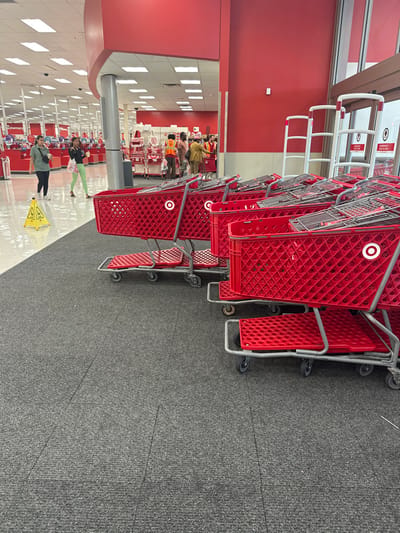 This image depicts the interior of a Target retail store in Fort Myers, United States. The scene is captured from a slightly elevated position near the entrance/exit, looking into the main retail area. On the right, multiple rows of red shopping carts, each featuring the white Target bullseye logo, are nested and positioned on a dark gray carpeted floor.
To the left, the flooring transitions to a light-colored tile, where a yellow, triangular "Caution Wet Floor" sign is visible. Two adult females are walking on this tiled area. The individual closer to the left wears a dark top and dark pants, carrying a dark shoulder bag. The second individual, slightly behind and to the right, wears a dark top and light green leggings, holding a white cup.
In the mid-ground to the right, a group of approximately five individuals is gathered near a service counter or entrance point. At least two individuals in this group are wearing orange safety vests over their clothing, while another wears a tan-colored outfit. The store interior features prominent red walls, red support columns, and a white ceiling equipped with rectangular fluorescent light fixtures. White security gate pedestals are visible near the store's exit point on the far right. Merchandise displays and additional store infrastructure are present in the background.