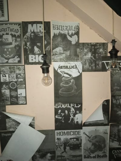 A wall covered with various black and white band posters dominates the scene. Notable bands include Buckethead, Mr. Big, Burzum, Metallica, and Soulfly. Other visible posters are Homicide, Burgerkill, Bats of Death and Cannibal Corpse. Two vintage-style light bulbs hang from the ceiling, adding a unique aesthetic to the display of musical artistry. The wall appears to be painted a light pink or peach color, contrasting with the dark posters.