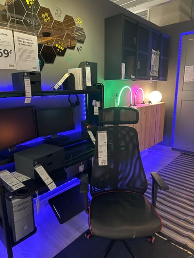The image shows an IKEA gaming room setup in Siero, Spain. The room features a black computer desk with two monitors, a keyboard, a desktop computer tower, and a black mesh gaming chair with red accents. Blue LED strip lights are installed under the desk and behind the monitors, creating a vibrant atmosphere.

On the wall behind the desk, there is a hexagonal mirror art piece with yellow gaming-themed icons, including a plus sign, a gamepad, and game-related text like "LVL UP" and "PLAY GAME". Above the desk, a dark grey cabinet with mesh doors is mounted on the wall. A poster for Iggy Pop's "Post Pop Depression Tour" is hanging on the cabinet.

To the right of the desk, a wooden cabinet with three doors is positioned against the wall. Two curved neon lights, one green and one pink, are placed on top of this cabinet, along with a spherical white lamp. The floor is covered with a light-colored wooden laminate and a striped rug.

Several price tags are visible throughout the scene, indicating that this is a retail display. For example, a tag on the gaming chair reads "189€". Other tags mention prices like "7,99€" and "12,99€". The overall impression is a modern, digitally-inspired gaming environment designed for comfort and style.