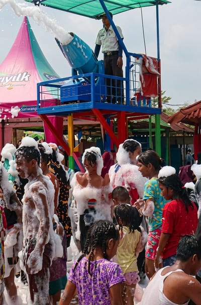 A scene at Durin Simbelang, Indonesia, features a group of people, including children, covered in foam, likely from a foam party. A man in uniform stands atop a blue platform operating a large, blue foam cannon, which is actively shooting foam onto the crowd below. Behind the platform, there's a pink and green tent with the word "Sariwangi" visible. The environment suggests an outdoor setting, possibly an amusement park or recreational area, indicated by the presence of other structures with colorful roofing and open areas. The people in the crowd are engaging with the foam, some looking amused or interacting with each other.