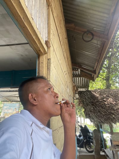 A man is in the foreground, his profile visible as he looks upwards and to the right, holding a lit cigarette to his lips. He wears a white collared shirt. Behind him, a weathered, yellow wooden building is prominent, with a corrugated metal roof extending over a thatched-roof structure. A dark-colored scooter is parked to the right, partially visible under the thatched roof. The background shows lush green foliage and trees, indicating an outdoor setting. A sign with text in an unfamiliar script is visible on the left side of the frame. The lighting suggests a sunny day. The scene appears to be in a rural or semi-rural location, possibly Meuriya, Indonesia, based on the context provided.