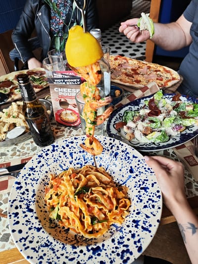 The image shows a table at a restaurant in Enderby, United Kingdom, laden with various food dishes and drinks. A person in a black leather jacket and floral top is seated at the table. Another person is seen holding a fork with salad.
The table features several dishes, including:
- A plate of pasta with tomato sauce and greens
- A platter of fried calamari with a dipping sauce
- A rectangular pizza topped with meat and vegetables
- A salad with lettuce, croutons, and bacon
- A shrimp skewer with a lemon on top, served in a glass
Other items present on the table include:
- A bottle of Gran Peroni beer
- Restaurant menus
- Silverware
- A decorative paper placemat
The table has a mosaic tile top. The overall scene suggests a casual dining experience.