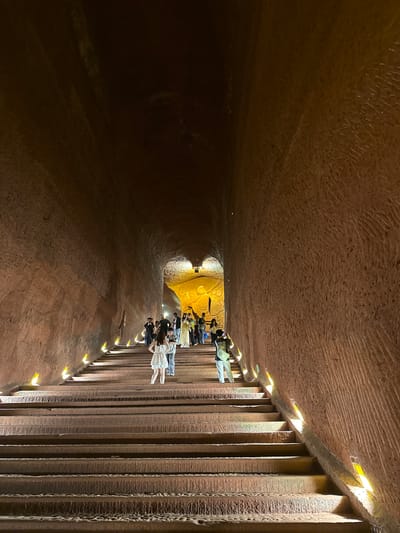 The image shows the Anyue Grottoes, located in Shizhong, China. The photo captures a group of people near the end of a tunnel, illuminated by artificial lighting. The tunnel appears to be carved from stone or earth. The walls have a textured, carved appearance. A row of vertical columns with built-in lights lines one side of the tunnel. The tunnel narrows as it extends into the distance.