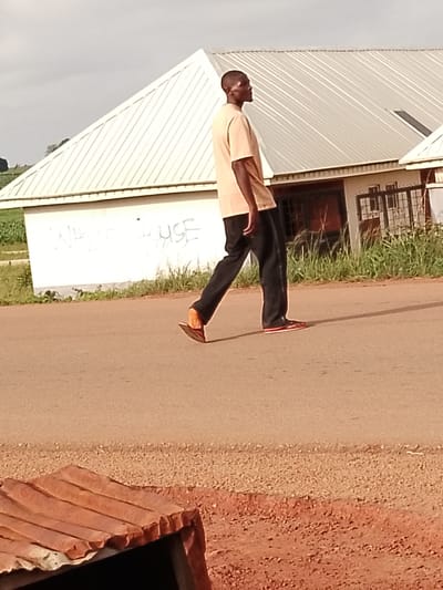 The image shows a man walking on a paved road in Agwala, Nigeria. He is wearing a light tan t-shirt and black pants, with a pair of sandals. The man is walking away from the camera, facing to the right. In the background, there is a building with a light-colored, corrugated metal roof and white walls. Graffiti reading "WHITE HOUSE" is visible on the wall. Tall green grass grows in front of the building and along the side of the road. The ground in the foreground is a reddish-brown dirt with scattered pebbles, and a corrugated metal roof of a structure is visible in the bottom left corner. The lighting suggests it is daytime, possibly late afternoon, with some shadows cast on the road. The sky is overcast.