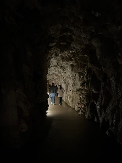 Two people stand on a brightly lit path within a dark cave. The cave's rough, rocky walls and ceiling surround them, contrasting sharply with the smooth path they occupy. The light source appears to originate from the path itself, illuminating their figures and the nearby cave walls. The rest of the cave is shrouded in darkness, creating a sense of mystery.