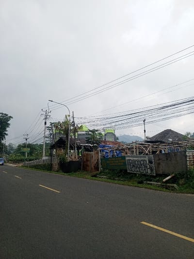 The scene depicts a roadside view under an overcast, light grey sky. An asphalt road, marked with two parallel dashed yellow lines, occupies the foreground and extends into the mid-ground, where a dark blue vehicle is partially visible on the far left, moving away from the viewer. On the right side of the road, multiple utility poles support numerous black electrical and communication cables strung horizontally across the frame. A street light with an attached lamp extends over the road from one of these poles.
Behind the poles and cables, a series of structures and dense green vegetation are present. A significant building with a prominent green roof and facade is visible further back. Closer to the road, there is a smaller, dark-roofed structure and a larger, temporary construction of wooden scaffolding draped with blue tarpaulins. A fence of rusty corrugated metal sheets stands between the road and these structures. Adjacent to the road, a green sign with white text is displayed, reading "PIMPINAN DAERAH PERHIMPUNAN ISLAM (PERSIS) KABUPATEN CIANJUR," indicating the location is within Cianjur Regency, Indonesia. Another rectangular panel, possibly concrete, featuring white graffiti, is positioned closer to the road. Hazy hills are discernible in the distant background. No people are visible within the scene, and no discernible actions or interactions are depicted.