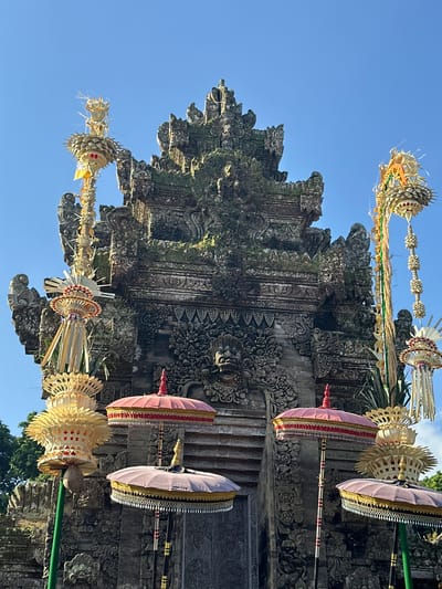 This is a front-facing, eye-level, medium shot of the Pura Kehen temple in Bangli, Indonesia, on a clear, sunny day. The temple's ornate stone gate, adorned with intricate carvings and moss, dominates the background. In the foreground, traditional Balinese offerings, known as *canang sari*, and decorative umbrellas are arranged. These offerings are made of woven leaves and adorned with yellow and red accents, while the umbrellas are layered with fabric and tassels in red, pink, and gold. The scene is devoid of people, and the focus is on the architecture and decorative elements of the temple. The sky is a vibrant blue, with no clouds visible.