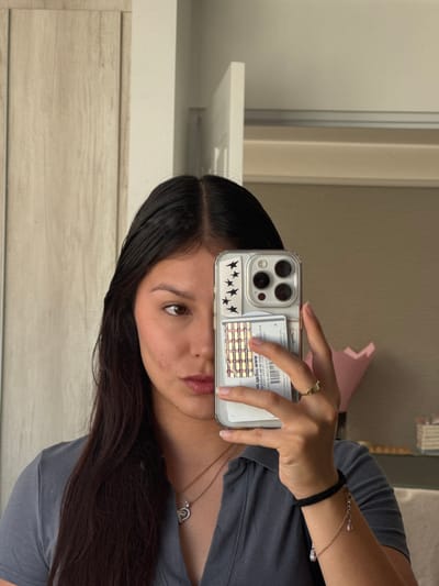A woman is taking a selfie with her phone. The phone has a clear case with black star stickers on the bottom and a credit card or ID tucked behind it. She is wearing a gray t-shirt, a necklace with a heart-shaped pendant, a ring, and a bracelet. She has long dark hair. The background includes a wall, a white door, and a glimpse of something pink. The OCR text on the phone case indicates web addresses.