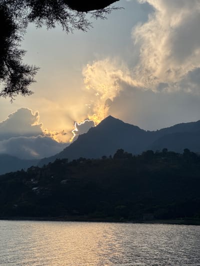 A landscape view features a body of water reflecting golden light on the right side. Mountain silhouettes dominate the center, contrasting sharply with the bright, sunlit clouds behind. The sun peeks through a gap, creating radiant beams. Silhouetted tree branches frame the lower left, adding depth to the scene. The overall mood is peaceful and serene.