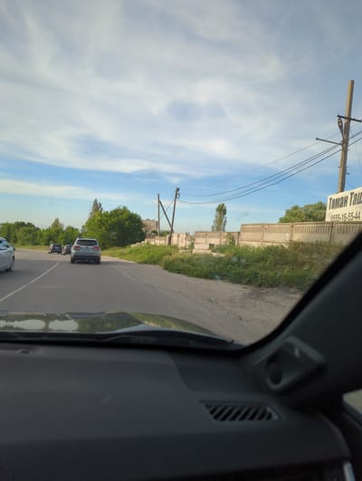 The image, apparently taken from inside a vehicle, depicts a road in Beş-Küŋgöy, Kyrgyzstan. The road is lined with vegetation on one side and a concrete wall with a sign in Russian on the other. Power lines run parallel to the road. Several vehicles are visible on the road, including at least one SUV. The sky is blue with scattered clouds. The interior of the vehicle from which the photo was taken is visible on the right side of the frame.