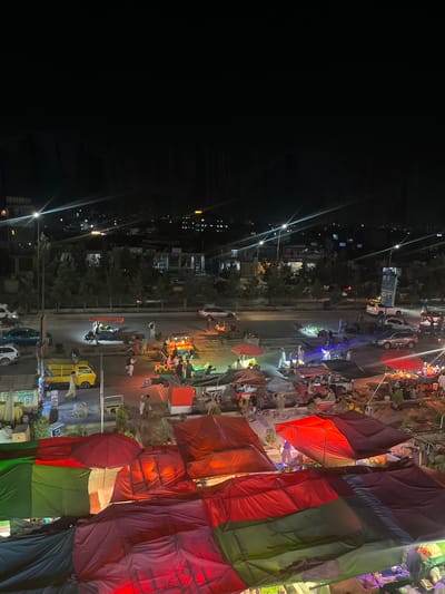 The image depicts a busy street scene at night in Kabul, Afghanistan. The foreground is dominated by a collection of outdoor stalls, many covered by colorful tarps and umbrellas in red, green, and dark shades. These appear to be food or goods vendors, with some displaying produce and various items.

Further back, across a paved road with streetlights, is another row of stalls and a street market area. Several vehicles, including cars and a yellow truck, are visible on the road. Pedestrians are present, walking around the stalls and interacting with vendors. Some individuals are gathered around the market stalls, suggesting active commerce.

In the background, the city of Kabul is illuminated by streetlights and building lights. Hills or mountains can be faintly seen against the dark night sky. The overall impression is a lively night market or street commerce area in an urban environment. The tarps in the foreground, featuring colors that resemble the Afghan flag (black, red, and green), suggest a patriotic or cultural theme to the market setup.