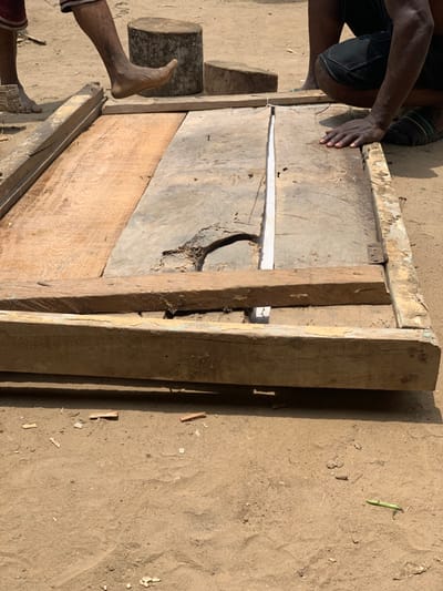 The image shows two people working on a wooden structure on a dirt ground in Egbeda, Nigeria. The structure is a large wooden frame made of several planks, which appear to be being joined together. One person is kneeling and using their hands to adjust the planks, while the other person's foot is resting on a large piece of wood that is propped against the structure. Small pieces of wood and sawdust are scattered around the structure on the ground. The scene is outdoors and the ground is sandy. The location based on the coordinates provided is in the suburb of Egbeda in Lagos, Nigeria.
