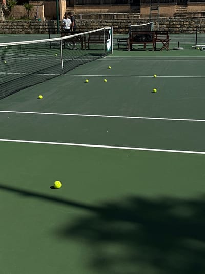 A forensic analysis reveals an outdoor tennis court situated in Ericeira, Portugal. The court features a dark green hard surface demarcated by white lines and a black mesh net with a white top tape and a green and white branded side strap on the right. Eleven yellow tennis balls are visible across the court, scattered on both sides of the net, with one ball prominent in the lower foreground. In the background, two individuals stand behind the net; one wears a white shirt and dark trousers, the other a dark shirt and dark trousers. Behind them, a stone retaining wall supports a level ground featuring two dark wooden benches and portions of a light-brown, possibly terracotta-colored, building. Strong sunlight illuminates the scene, creating sharp shadows from the net, balls, and other unseen elements to the lower right.